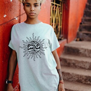 May include: A person wearing a light blue t-shirt with a black and white sun graphic. The person is standing in front of a red wall with white writing.