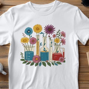May include: White t-shirt featuring a colorful floral design. The graphic shows flowers in laboratory beakers and flasks. The flowers are yellow, pink, purple, and blue. The beakers and flasks have blue and yellow liquid.