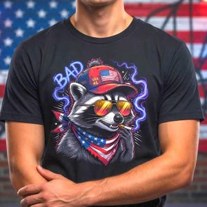 May include: Black t-shirt featuring a raccoon wearing sunglasses, a USA #1 hat, and a bandana with an American flag design. The word "BAD" is written above the raccoon in blue. The background includes an American flag.