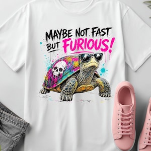 May include: White t-shirt featuring a colorful turtle wearing sunglasses with the text "MAYBE NOT FAST BUT FURIOUS!". The turtle has a skull design on its shell. Pink sneakers and gray jeans are also in the image.