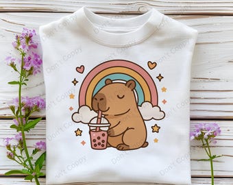 May include: White t-shirt with a cartoon capybara drinking bubble tea in front of a rainbow. The capybara is brown and has a pink nose and closed eyes. The bubble tea is in a clear cup with a brown lid and a straw.