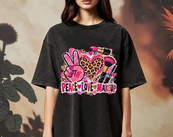 Może przedstawiać: Czarny t-shirt z efektem sprania, ozdobiony grafiką przedstawiającą różowy znak pokoju, serce w panterkę, szminkę, pędzel do makijażu i tusz do rzęs. Poniżej znajduje się napis PEACE LOVE MAKEUP wykonany różowymi literami.