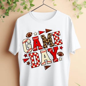 May include: White t-shirt with the words "GAME DAY" in a colorful design. The letters are in red, beige, and leopard print patterns, with footballs, baseballs, and stars. The shirt is hanging on a black hanger.