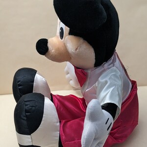 May include: A plush Mickey Mouse toy wearing a white tuxedo with a red bow tie and red pants. The toy is sitting with its legs crossed and has a black and white design on its shoes.