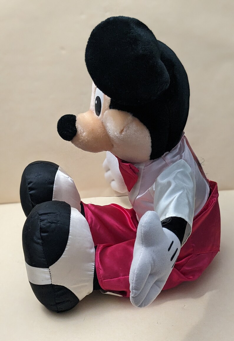 May include: A plush Mickey Mouse toy wearing a white tuxedo with a red bow tie and red pants. The toy is sitting with its legs crossed and has a black and white design on its shoes.