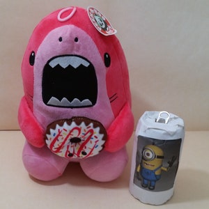 May include: Pink plush shark toy holding a pink and white donut with sprinkles. The shark has a large open mouth with white teeth. The toy has a small tag attached to its head that says "MOOO!"