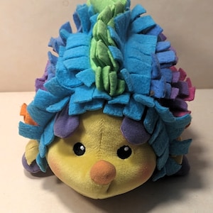 May include: A colorful plush toy hedgehog with a yellow face, blue, green, purple, and orange fur. The toy has a textured, fuzzy surface.