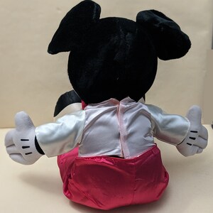 May include: A plush toy of Mickey Mouse wearing a white shirt with a pink satin bow tie and pink satin pants. The toy is sitting with its arms outstretched.