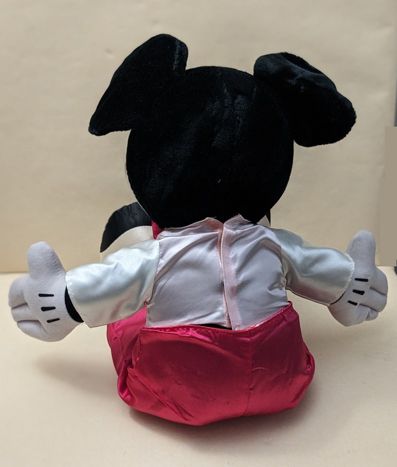 May include: A plush toy of Mickey Mouse wearing a white shirt with a pink satin bow tie and pink satin pants. The toy is sitting with its arms outstretched.