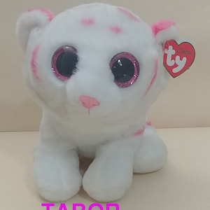 Puede incluir: Tigre de peluche blanco con rayas rosas y ojos con purpurina rosa. El tigre está sentado con las patas metidas debajo del cuerpo. La etiqueta dice "Ty Classic".