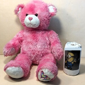 May include: Pink plush teddy bear with white accents and a heart design on one paw. The bear is sitting on a white surface.