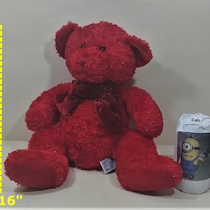 Puede incluir: Un oso de peluche rojo, de unos 40 cm de altura, con un lazo de terciopelo a juego. El oso tiene una textura brillante y está junto a un pequeño recipiente cilíndrico con un personaje de Minion impreso.
