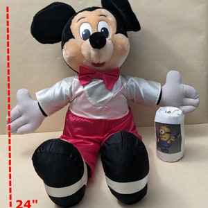 May include: A large plush Mickey Mouse toy wearing a red bow tie and red pants. The toy is sitting down with its arms outstretched. The toy is 24 inches tall.