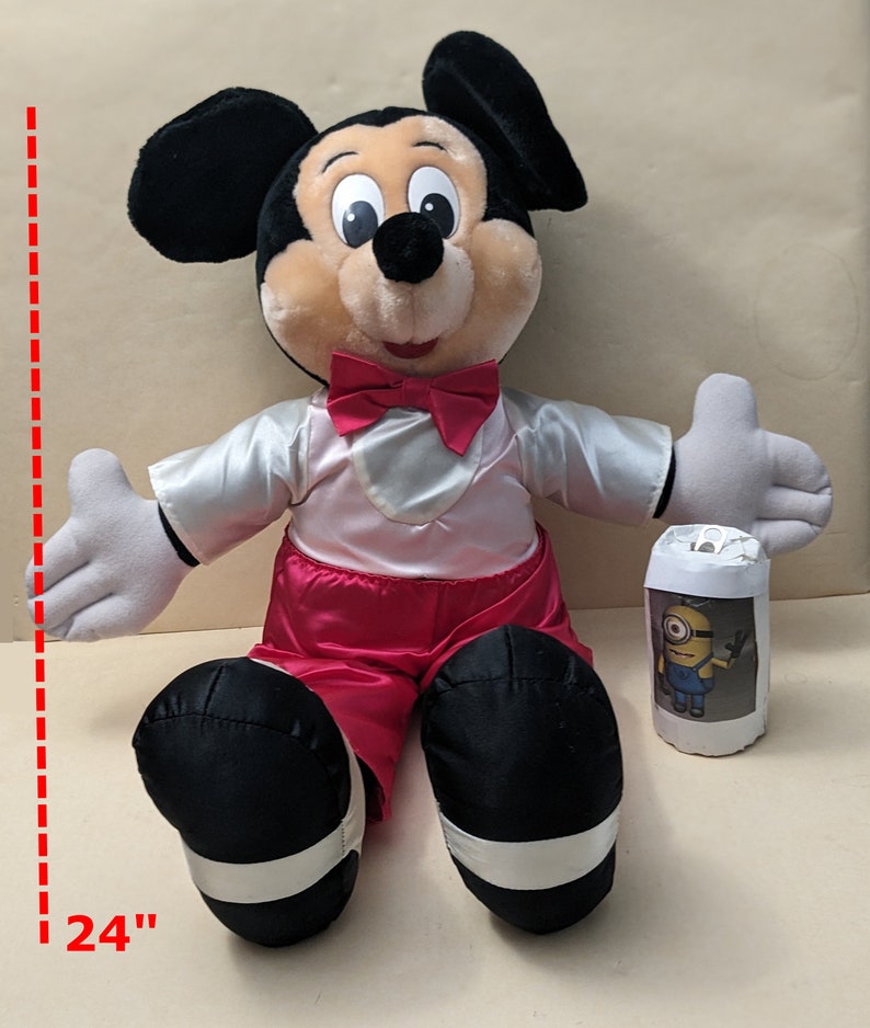 May include: A large plush Mickey Mouse toy wearing a red bow tie and red pants. The toy is sitting down with its arms outstretched. The toy is 24 inches tall.