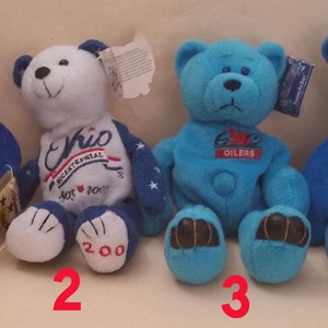 Puede incluir: Cuatro osos de peluche, cada uno con un uniforme diferente. El primer oso lleva un uniforme azul con una corbata de lazo blanca y un parche azul marino con un águila blanca. El segundo oso lleva un uniforme blanco y azul con la palabra "Ohio" en la parte delantera. El tercer oso lleva un uniforme azul con la palabra "Oilers" en la parte delantera. El cuarto oso lleva un uniforme azul con el número "98" en la parte delantera.