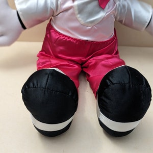May include: A plush toy with white gloves, a pink shirt, and red pants. The toy is wearing black shoes with white trim.