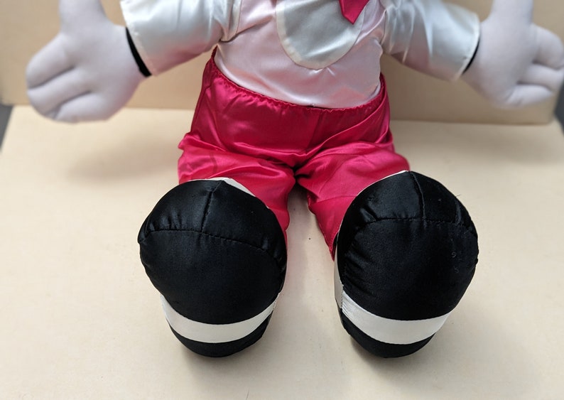 May include: A plush toy with white gloves, a pink shirt, and red pants. The toy is wearing black shoes with white trim.