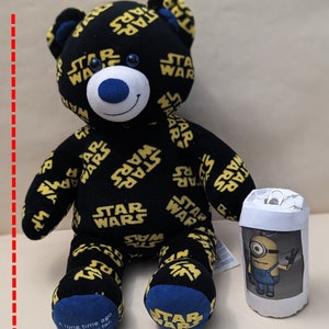 May include: A black plush bear, approximately 43 cm tall, with yellow "STAR WARS" text. The bear has blue accents on the ears, paws, and nose. A small, cylindrical container with a Minion graphic is next to the bear.