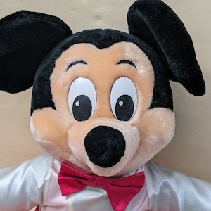 May include: A plush Mickey Mouse toy wearing a white shirt with a red bow tie. The toy has black ears, a black nose, and large white eyes with black pupils.
