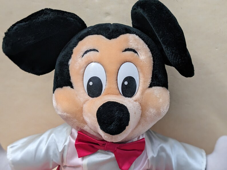 May include: A plush Mickey Mouse toy wearing a white shirt with a red bow tie. The toy has black ears, a black nose, and large white eyes with black pupils.