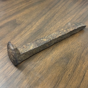 May include: A rusty metal railroad spike with a blunt head and a pointed end.