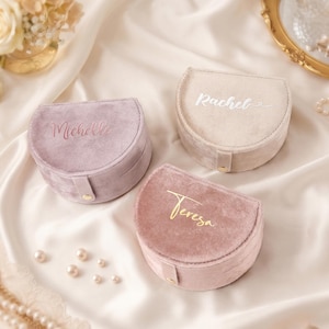Op de afbeelding: Drie fluwelen sieradendoosjes in roze en beige tinten, elk met een naam in elegant schrift. De doosjes zijn halfrond met een gouden sluiting. De achtergrond is een zachte, witte stof met parels en bloemen.