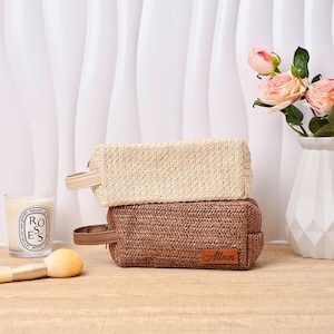 Puede incluir: Dos neceseres rectangulares tejidos, uno beige y otro marrón, apilados sobre una superficie de madera. La bolsa marrón tiene una etiqueta de cuero con el nombre "Allison". También hay una vela, un pincel de maquillaje y un jarrón con rosas rosas.