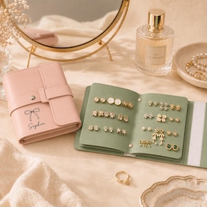 May include: A light pink leather jewelry organizer with a snap closure and the name Sophia printed above a bow graphic. Beside it, an open sage green jewelry book displays various stud earrings, including floral, star, and bow-shaped designs.