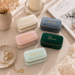 Op de afbeelding: Een verzameling kleine, rechthoekige sieradendoosjes in verschillende kleuren: crème, blauw, roze, groen en mint. Elk doosje heeft een gouden rits en een met goud geborduurde naam. De doosjes zijn op een neutrale ondergrond geplaatst.