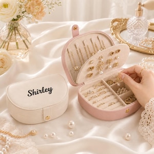May include: A cream-coloured jewellery box with the name Shirley printed in black, paired with an open pink jewellery box containing various gold rings, earrings, and necklaces. The interior features velvet-lined compartments and hooks for organised storage.