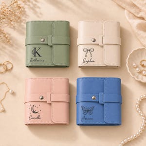 May include: Four square wallets with snap-button straps in sage green, cream, dusty pink, and blue. Each wallet features a unique black graphic and name: Katherine with a letter K, Sophia with a bow, Camille with a crescent moon, and Ariana with a butterfly.