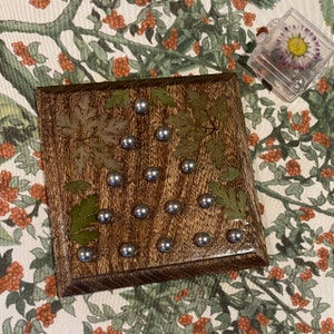 May include: A wooden game board with a dark brown stain. The board has 15 silver metal pegs and is decorated with pressed green leaves.