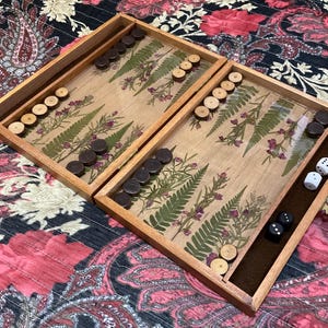 May include: A wooden backgammon set with a natural wood finish. The game board features a decorative design with pressed ferns and small purple flowers. The set includes brown and light-colored game pieces, and dice.