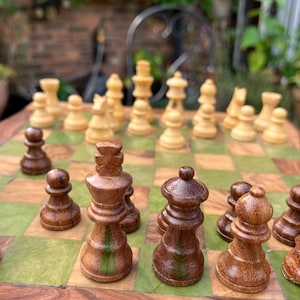 May include: A wooden chess set with green and brown squares. The chess pieces are made of wood and have a natural finish. The pieces are arranged on the board in a game in progress. The king and queen pieces are in the foreground.