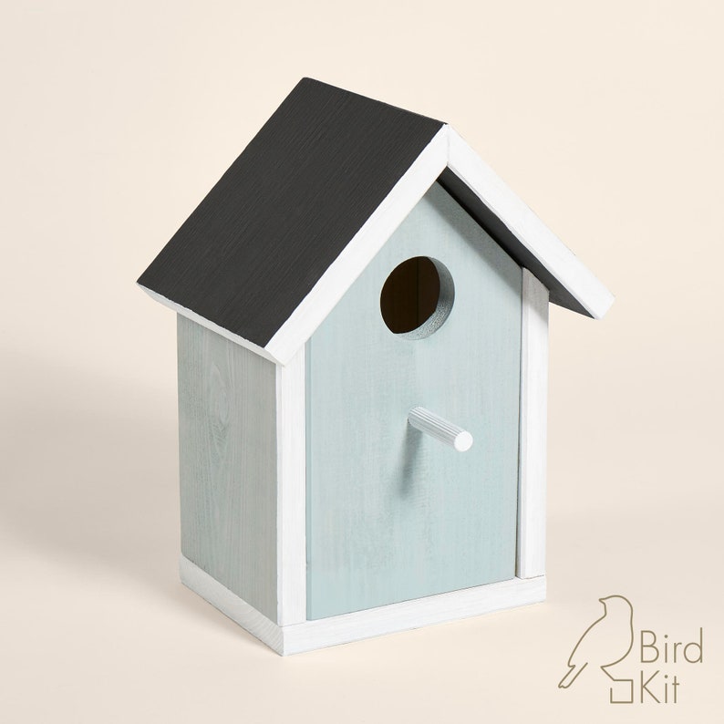 K&ouml;nnte beinhalten: Ein hellblaues Vogelhaus mit schwarzem Dach und wei&szlig;en Verzierungen. Das Vogelhaus hat ein rundes Eingangsloch und einen kleinen Sitzplatz.