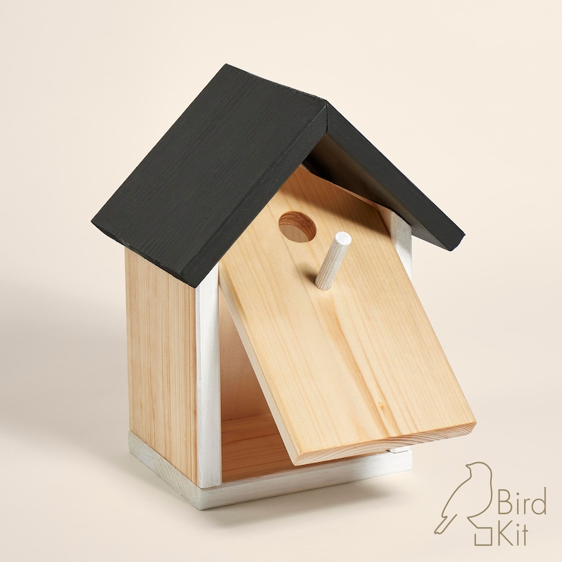 K&ouml;nnte beinhalten: Ein Vogelhaus aus Holz mit einem schwarzen Dach und wei&szlig;en Verzierungen. Die Vorderseite des Hauses hat eine Fl&uuml;gelt&uuml;r, die ge&ouml;ffnet ist und den Innenraum zeigt. Es gibt ein kleines rundes Loch an der Vorderseite des Hauses, durch das V&ouml;gel eintreten k&ouml;nnen.