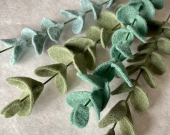 May include: Three stems of felt eucalyptus leaves in shades of green and blue. The leaves are arranged in a cascading pattern.
