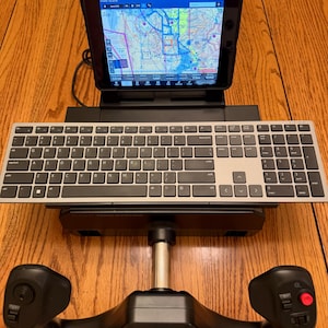 May include: A black flight simulator yoke system with a silver and black wireless keyboard mounted above it. A tablet is positioned behind the keyboard, displaying a digital aeronautical navigation map with various flight data and interface icons.