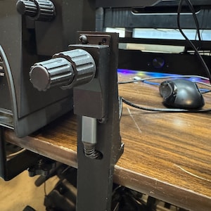 May include: A black metal desk clamp with two knobs. The clamp is attached to a desk and has a gray cable running through it.