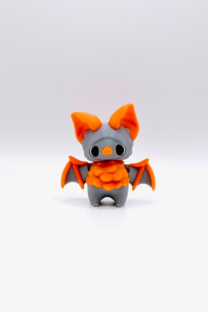 Peut inclure: Une petite figurine de chauve-souris en plastique gris imprim&eacute;e en 3D avec des ailes orange et un corps orange duveteux.