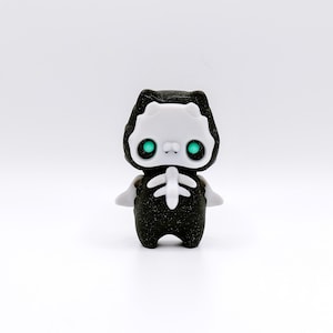 May include: A small black and white figurine of a cat wearing a skeleton costume. The cat has green eyes and a sparkly black body.