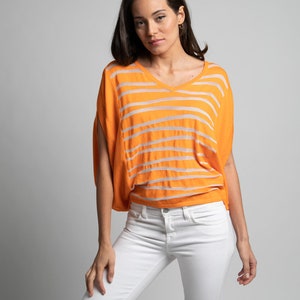 Puede incluir: Una mujer con una camisa naranja y blanca a rayas con cuello en V y pantalones vaqueros blancos.