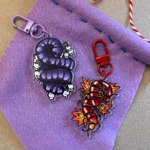 May include: Two keychains with metal clasps. One features a purple caterpillar design with white skull accents. The other has a red centipede design with orange and yellow flame accents. Both are on a purple felt pouch.
