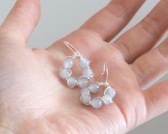 May include: A pair of hoop earrings with pale gray beads. The beads are strung on silver wire, forming a circular shape. The earrings are displayed on a light-colored background.