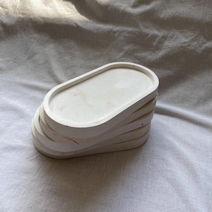 Pode incluir: Um conjunto de quatro bandejas ovais de concreto com acabamento branco e veios sutis. As bandejas estão empilhadas umas sobre as outras, sendo a bandeja superior a maior e a bandeja inferior a menor.
