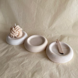 Op de afbeelding: Drie witte keramische schalen, twee ronde en een ovale, met een witte touwkaars in de middelste schaal. De ovale schaal bevat een houten stok.
