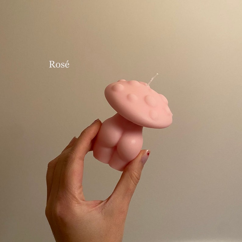 K&ouml;nnte beinhalten: Eine rosa Kerze in Form eines Pilzes mit einer runden Oberseite und einer Unterseite, die einem menschlichen K&ouml;rper &auml;hnelt. Die Kerze hat einen wei&szlig;en Docht.