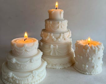 May include: Three white wedding cake candles, each with a lit flame in the center. The largest candle has four tiers, the middle candle has three tiers, and the smallest candle has two tiers. All three candles are decorated with white frosting and intricate details.