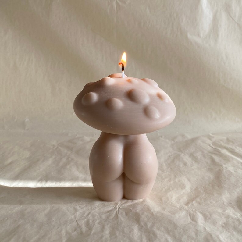 K&ouml;nnte beinhalten: Eine nude-farbene Kerze in Form eines Pilzes mit einem brennenden Docht. Die Oberseite der Kerze ist wie ein Pilzhut geformt mit einer strukturierten Oberfl&auml;che.