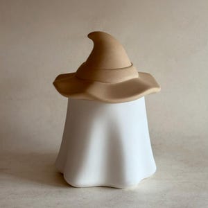 Puede incluir: Decoración de Halloween de cerámica con una figura de fantasma blanco que lleva un sombrero de bruja marrón. El sombrero tiene una corona puntiaguda y un ala ancha. El fantasma tiene una forma redondeada y fluida.